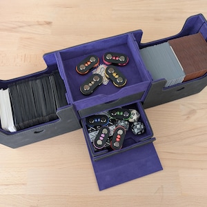 May include: A multi-compartment card game storage box in black and purple. The box is open, revealing stacks of cards, dice, and game accessories. The interior is lined with purple fabric, and the exterior is black.