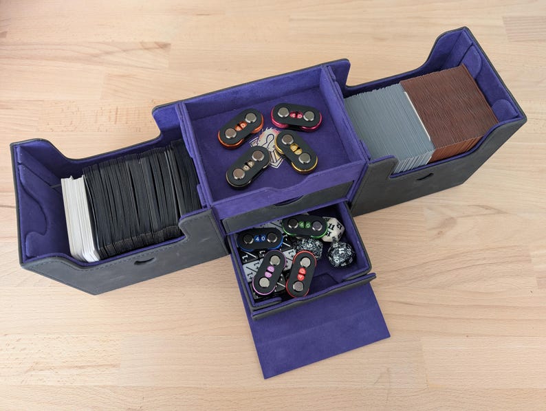 May include: A multi-compartment card game storage box in black and purple. The box is open, revealing stacks of cards, dice, and game accessories. The interior is lined with purple fabric, and the exterior is black.