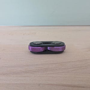 May include: A black key organizer with two silver circular accents and two purple, ridged wheels. The key organizer is resting on a light brown wooden surface, with a light blue wall in the background.