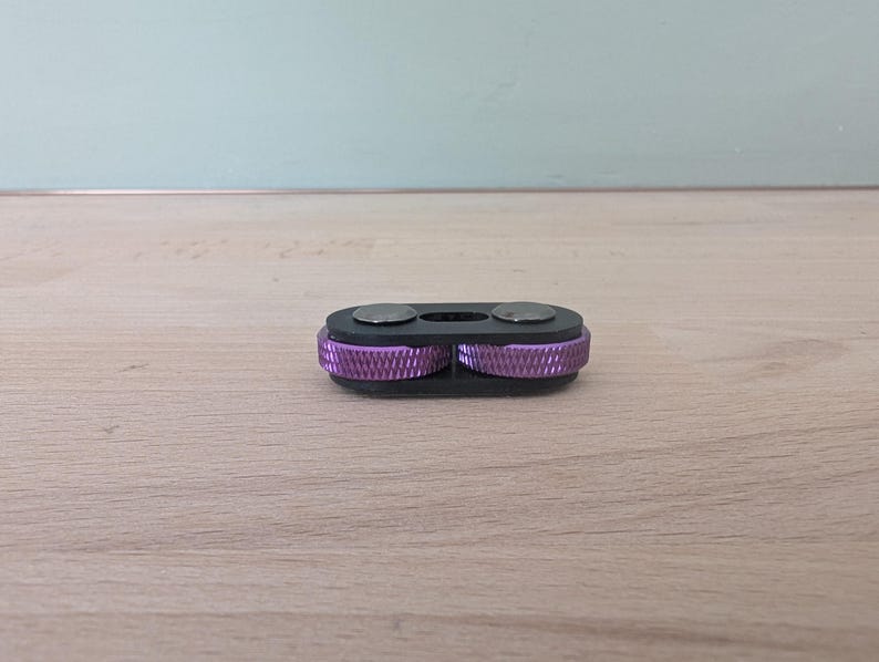 May include: A black key organizer with two silver circular accents and two purple, ridged wheels. The key organizer is resting on a light brown wooden surface, with a light blue wall in the background.