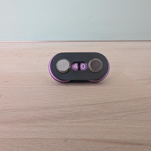 May include: A small, black and purple combination lock with two circular dials and a digital display showing the number "40". The lock has a rounded rectangular shape and is set against a light wood surface.