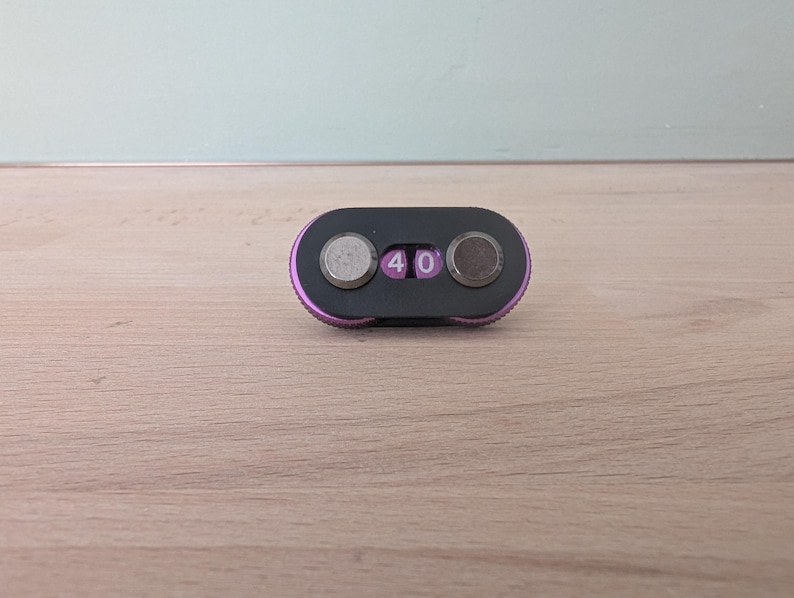 May include: A small, black and purple combination lock with two circular dials and a digital display showing the number "40". The lock has a rounded rectangular shape and is set against a light wood surface.