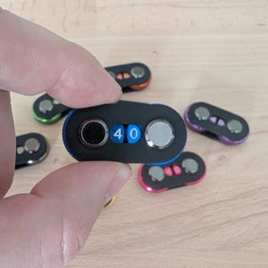 May include: A black combination lock with the number 40 displayed. The lock is held in a hand, with several other locks of different colors in the background. The locks have a rounded, oval shape with two dials and a small display.