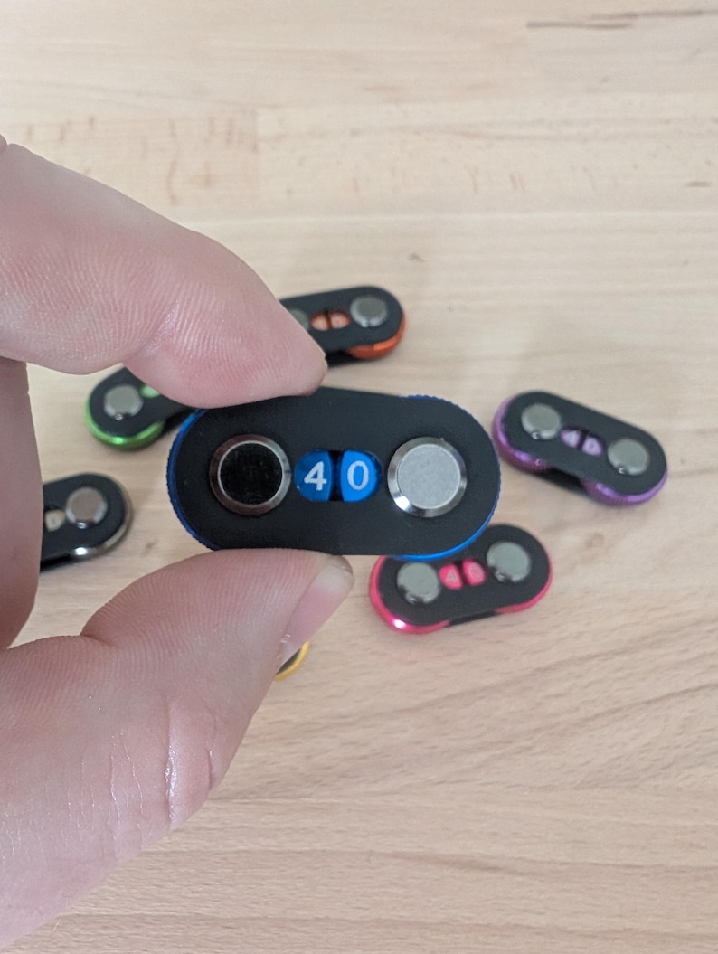 May include: A black combination lock with the number 40 displayed. The lock is held in a hand, with several other locks of different colors in the background. The locks have a rounded, oval shape with two dials and a small display.
