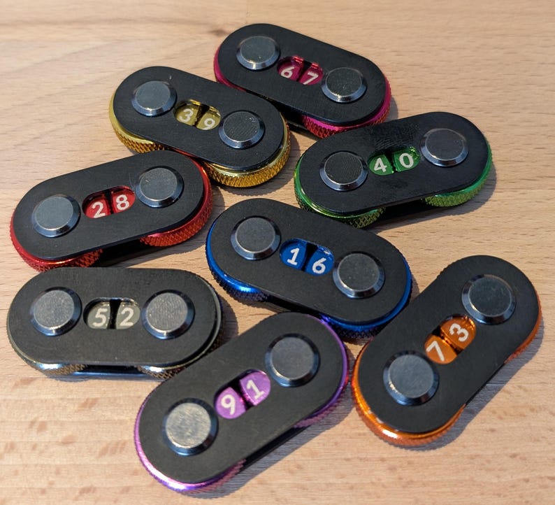 May include: A collection of black oval-shaped combination locks with metallic accents and colored rims. Each lock displays a unique two-digit number in white or orange. The rims are in various colors, including red, gold, green, blue, purple, and orange.