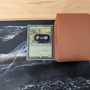 May include: A Magic: The Gathering card, "Chatterfang, Squirrel General", with a black and blue counter. A stack of brown card sleeves sits to the right. The card rests on a black and white playmat with a swirling design.