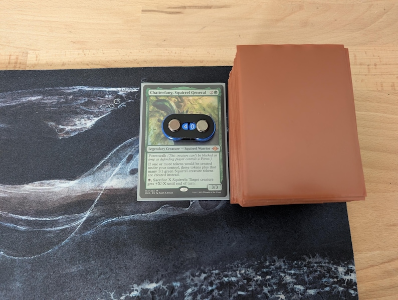 May include: A Magic: The Gathering card, "Chatterfang, Squirrel General", with a black and blue counter. A stack of brown card sleeves sits to the right. The card rests on a black and white playmat with a swirling design.