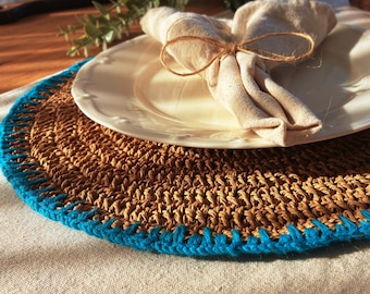 Può includere: Un sottopiatto marrone intrecciato con un bordo blu brillante, sormontato da un piatto bianco e un tovagliolo beige piegato legato con spago. Il sottopiatto è su una tovaglia chiara, creando un'atmosfera rustica.