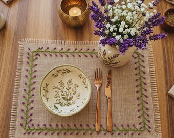Può includere: Un'apparecchiatura rustica con un sottopiatto beige con un bordo ricamato verde e viola. Una ciotola in ceramica con un design botanico, posate color rame e un vaso di fiori di lavanda e bianchi sono disposti. Una candela accesa in un portacandele dorato.