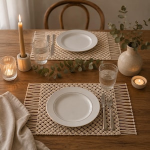 May include: A dining table setting with two white plates, macrame placemats, glasses of water, silverware, a candle, and a vase with greenery. The table is made of wood, and the scene is lit with natural light.