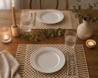 Può includere: Tavola apparecchiata con due piatti bianchi, tovagliette in macramè, bicchieri d'acqua, posate, una candela e un vaso con vegetazione. La scena è illuminata dalla luce naturale, creando un'atmosfera calda.