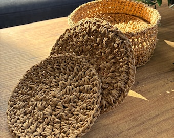 Può includere: Un cestino intrecciato con due sottobicchieri abbinati. Il cestino è rotondo e aperto, con un aspetto marrone chiaro strutturato. I sottobicchieri sono anch'essi rotondi e presentano un design intrecciato simile. Gli articoli sono in materiali naturali.