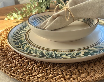 Può includere: Un'apparecchiatura da tavola con un sottobicchiere intrecciato, un piatto bianco con un motivo a foglie verdi e blu e una ciotola abbinata. Un tovagliolo di lino piegato è legato con spago. Perfetto per un pasto informale.
