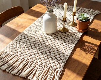 Può includere: Runner da tavolo in macramè color crema con frange, su un tavolo di legno. Il runner è decorato con un vaso bianco con lavanda, due candele accese in portacandele in ottone e una piccola pianta grassa in vaso.