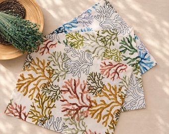 Può includere: Tre tovaglioli di stoffa quadrati con sfondo bianco e un motivo ripetuto di rami di corallo nelle tonalità giallo senape, verde salvia, terracotta e contorni neri. I tovaglioli sono impilati, mostrando la colorata stampa botanica.