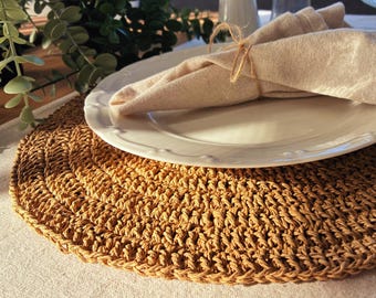Può includere: Un sottopiatto rotondo intrecciato di colore beige naturale, con un piatto bianco e un tovagliolo beige piegato. Il tovagliolo è legato con spago. Il sottopiatto è realizzato in un materiale strutturato.
