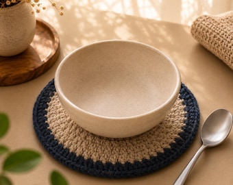 Può includere: Una ciotola in ceramica beige è appoggiata su un sottobicchiere rotondo all'uncinetto fatto a mano con un bordo blu navy. Un cucchiaio d'argento è appoggiato accanto al sottobicchiere. Un piccolo vaso con fiori secchi e un vassoio di legno sono sullo sfondo.