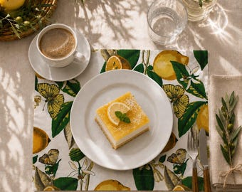 Può includere: Una tovaglietta quadrata con una stampa botanica di limoni gialli, foglie verdi e farfalle su sfondo bianco. Un piatto bianco contiene un dessert quadrato al limone con una fetta di limone e menta, accanto a una tazza di caffè.