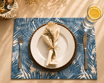 Può includere: Una tovaglietta rettangolare con motivo a foglie di palma blu e bianco, sormontata da un sottopiatto intrecciato, un piatto in ceramica bianca, un tovagliolo di stoffa beige legato con spago e rosmarino, forchetta, coltello e bicchiere d'acqua.