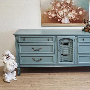 May include: A vintage-style, blue dresser with six drawers and a cabinet with a wire mesh door. The dresser has ornate hardware and sits on four legs. A white poodle statue is on the left, and a floral painting hangs above.