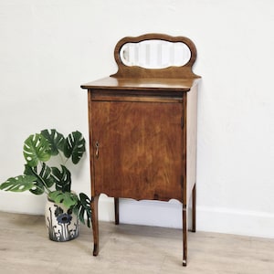 May include: A vintage wooden cabinet with a small mirror on top. The cabinet is a warm brown color with a single door and a keyhole. The legs are curved, and the mirror has a decorative shape. A potted plant is visible on the left.