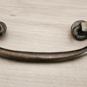 May include: A bronze-toned drawer pull with a curved handle and square mounting hardware. The hardware is attached to circular backplates. The pull is designed for furniture and cabinetry.