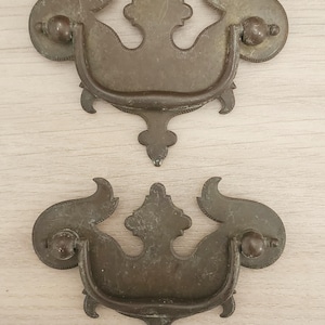 May include: Two antique bronze-toned drawer pulls. Each pull features a curved handle with decorative scrollwork and a central ornate design. The pulls have a vintage aesthetic, suggesting they are for furniture restoration or decorative projects.