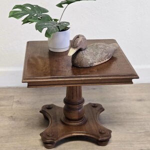 May include: A dark brown, square-top side table with a pedestal base. A decorative duck decoy and a potted plant with green leaves sit on the table. The table is made of wood and has a classic design.