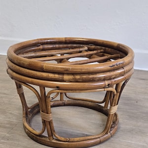 May include: A round, low-profile stool constructed from natural brown rattan. The piece features a circular top frame with horizontal slats and a base formed by curved rattan supports, all secured with light-colored binding at the joints.