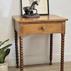 May include: A light brown wooden side table featuring a single drawer with a small round knob and four distinctive bobbin-style legs. The table has a flat rectangular top surface and a smooth finish, showcasing the natural grain of the wood.