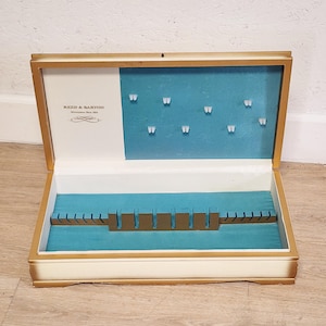 May include: A white wooden cutlery box with gold trim. The box is open and has a blue velvet lining. The box has a Reed & Barton logo and a label that says "Silversmiths Since 1824".