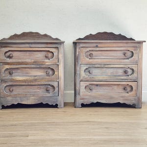May include: Two matching wooden nightstands with three drawers each. The nightstands have a distressed, weathered finish in shades of brown and gray. Each drawer has a decorative pull and the top has a scalloped edge.