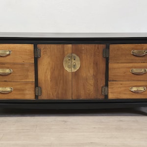 May include: A long rectangular sideboard featuring a black frame and six natural wood drawers with brass bail pulls. The center section has two cabinet doors with a circular brass plate and latch. The piece stands on four curved feet.
