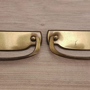 Puede incluir: Dos tiradores de cajón antiguos de latón, con una placa trasera rectangular y un asa curvada. Se encuentran sobre una superficie de madera marrón claro. El herraje tiene un aspecto vintage y envejecido.