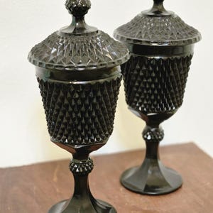 May include: Two black glass lidded compotes featuring a prominent diamond point pattern on the bowls and lids. Each piece stands on a pedestal base with a small decorative knop on the stem and a pointed finial on the top of the removable lid.