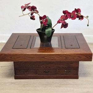 May include: A dark wood coffee table with three drawers and a rectangular top. The top features a central glass panel and two wooden panels. A black pot with burgundy flowers sits on top of the table.