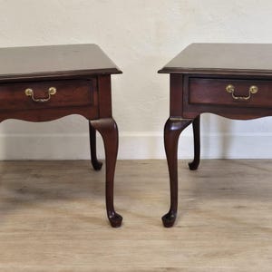May include: Two matching mahogany side tables. Each table has a square top, a single drawer with a brass pull, and curved legs. The tables are a rich, dark brown color and are set against a neutral background.