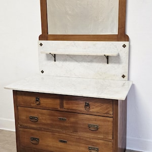 May include: A wooden dresser featuring a white marble top and a matching marble backsplash with a small shelf. The unit includes two small top drawers and two larger lower drawers, all fitted with metal ring pulls and central keyhole escutcheons.
