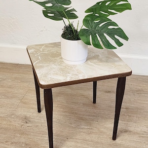 May include: A small, square side table with dark brown, tapered legs and a light marble-patterned top. A white, ribbed ceramic pot with a faux Monstera plant sits on the table. The plant has large, green, split leaves.