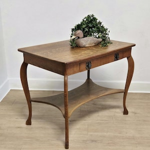 May include: A rectangular wooden side table with a medium brown finish, featuring a single drawer with two dark metal floral-shaped pulls, curved legs, and a lower shelf. A decorative wooden duck decoy sits atop the table with green foliage.