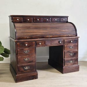 May include: A dark brown wooden roll-top desk featuring a slatted tambour door, a top row of six small drawers with white knobs, and a central workspace with one wide drawer. The base includes eight additional drawers with metal pulls and knobs.