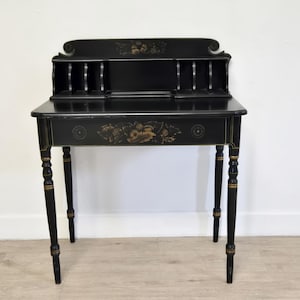 May include: Black antique writing desk with a decorative floral design and gold accents. The desk has a small hutch with vertical dividers and a single drawer. The legs are slender and have gold detailing. A classic piece of furniture.