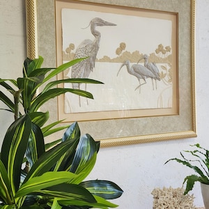 May include: A framed print of a grey heron and two white ibises standing in a marsh. The print is in a gold frame with a cream mat.