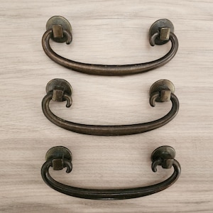 May include: Three antique bronze drawer pulls with a curved handle design. Each pull features a circular base with a decorative element and a curved handle. The hardware is displayed on a light wood grain surface.