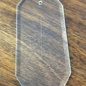 May include: Clear, geometric glass pendant with a small hole at the top. The pendant has a starburst design etched into the center. The pendant is on a wooden surface.