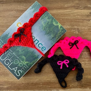 以下が含まれることがあります： 3つのクロシェ編みのランジェリー風ブックマークのデザイン。1つは黒のトリムが付いた赤、1つは黒のトリムが付いたピンク、もう1つはピンクのトリムが付いた黒。それぞれに黒いリボンが付いています。