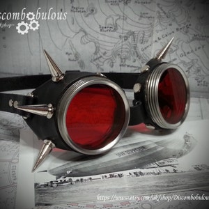 May include: A pair of steampunk goggles with red lenses and silver accents. The goggles are adorned with spikes and have a black leather strap.