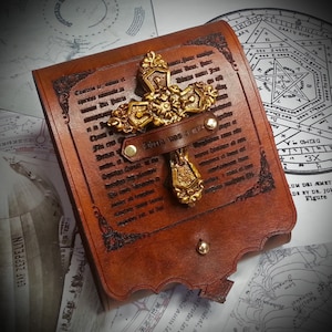 May include: A brown leather journal cover with a gold cross design and the text "Divini Nos Est" embossed on the front. The cover has a decorative border and metal rivets.