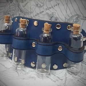 May include: A blue leather wristlet with four small glass bottles with cork stoppers. The wristlet has silver metal rivets and is designed to hold small vials.