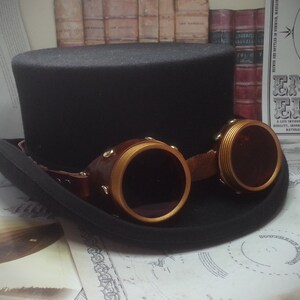 May include: A black top hat with a wide brim and a pair of gold-rimmed goggles with brown leather straps. The goggles have round lenses and are attached to the hat with a strap.